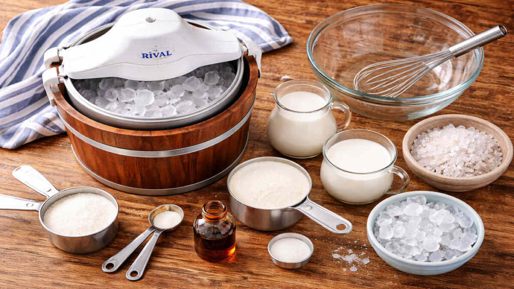 Rival ice cream maker with wooden bucket, milk, sugar, rock salt, whisk, and mixing bowls on a rustic wooden kitchen counter.