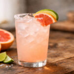 Paloma cocktail in a highball glass with ice, grapefruit slice, lime wedge, and salt rim on a rustic wooden table.